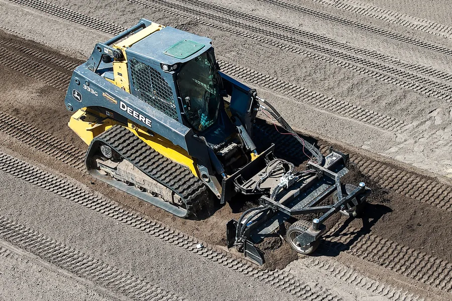 Aerial view of a John Deere 333G track loader pulling a power rake across a freshly graded lot
